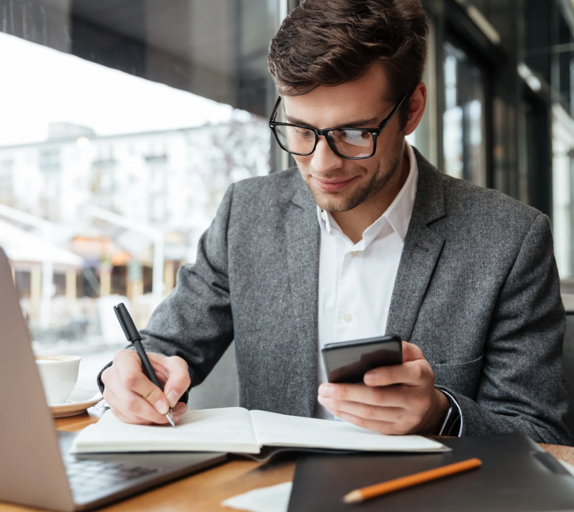 Young man in glasses and gray blazer writing in a notebook while holding a smartphone at a table with a laptop and coffee cup.