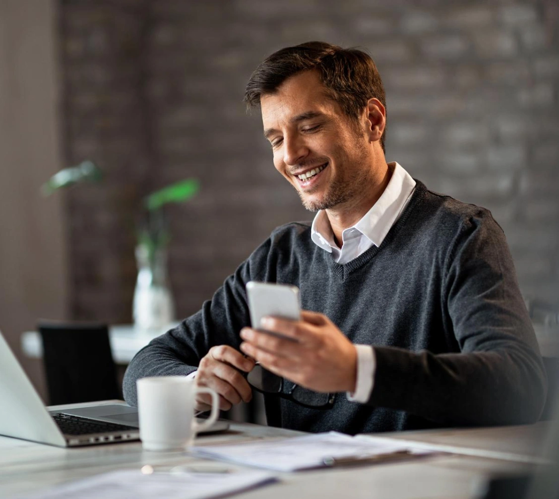 Smiling man in a dark sweater sitting at a desk with a laptop, holding a smartphone and glasses.