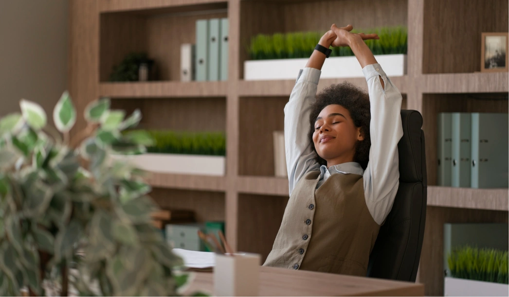 Woman sitting in office chair stretching with eyes closed and smiling, with shelves and green plants in background.