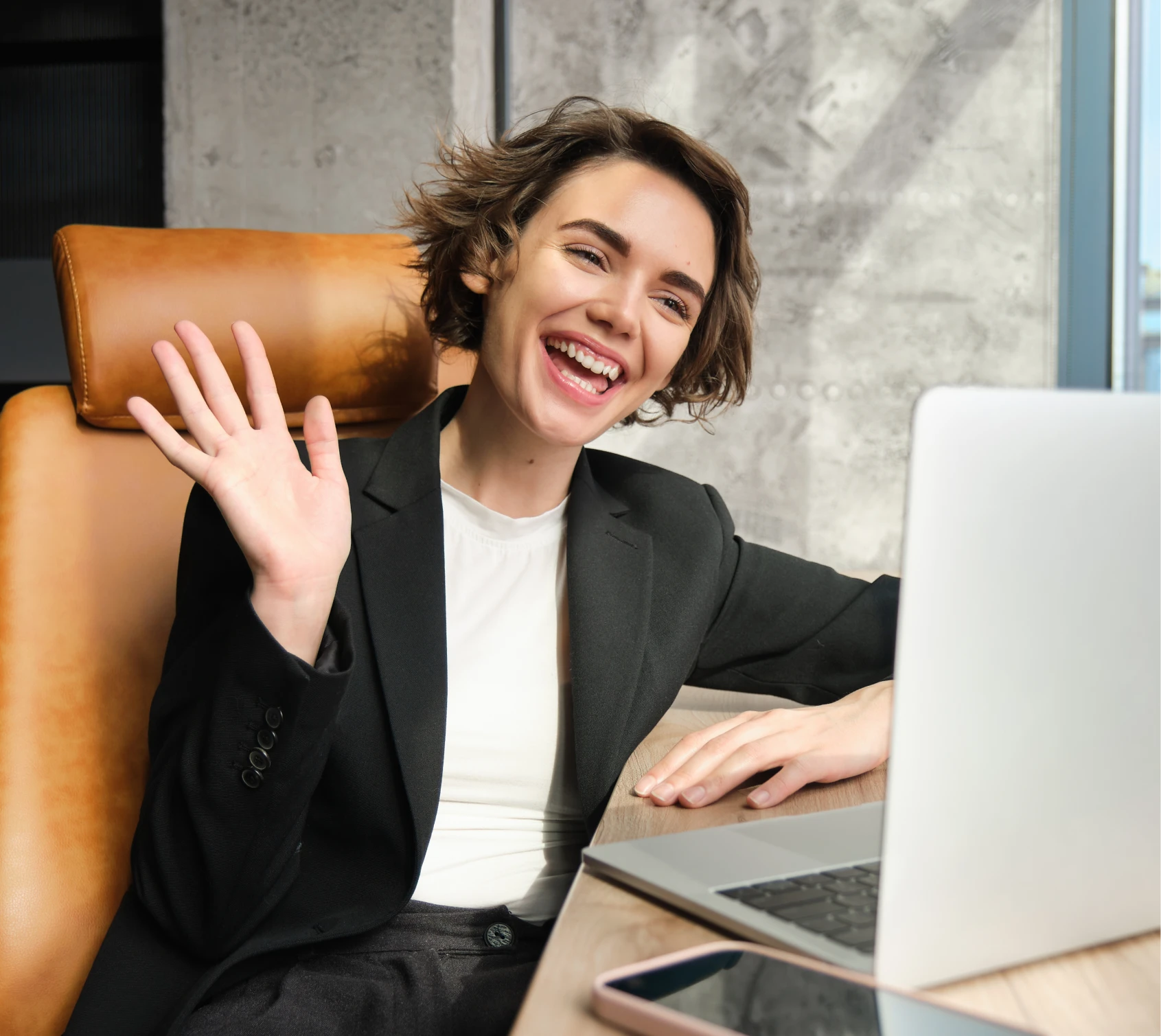 Smiling woman in a black blazer waving during a video call while sitting on a tan chair with a laptop and smartphone on the desk.