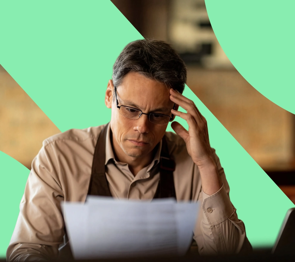Man wearing glasses and apron looking concerned while reading papers at a desk.