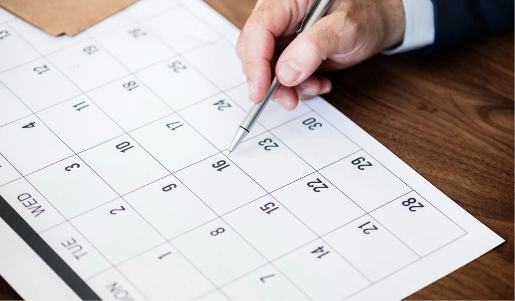 Close-up of a hand holding a pen pointing to the 16th date on a calendar.