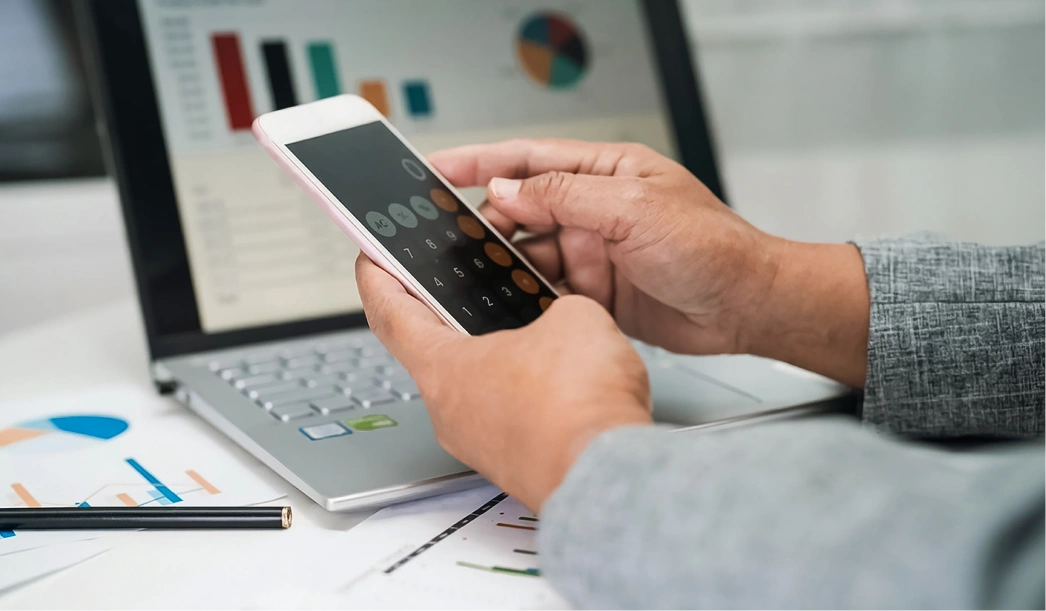 Person using a calculator app on a smartphone with financial charts and a laptop in the background.