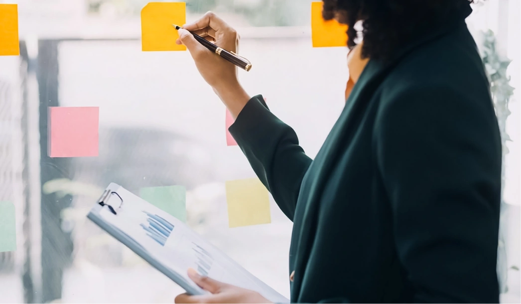 Person writing on orange sticky notes on a glass board while holding a clipboard with charts.