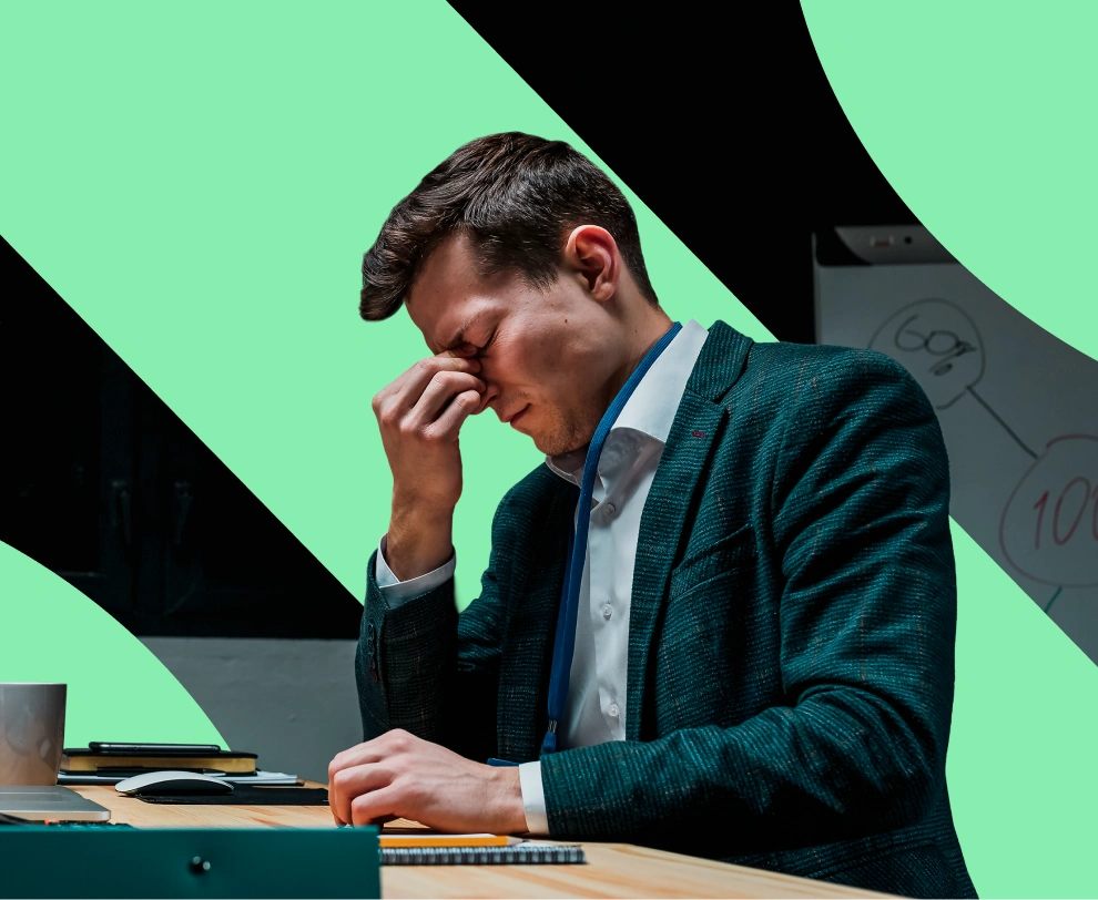 Stressed man in a suit sitting at a desk holding the bridge of his nose with eyes closed.