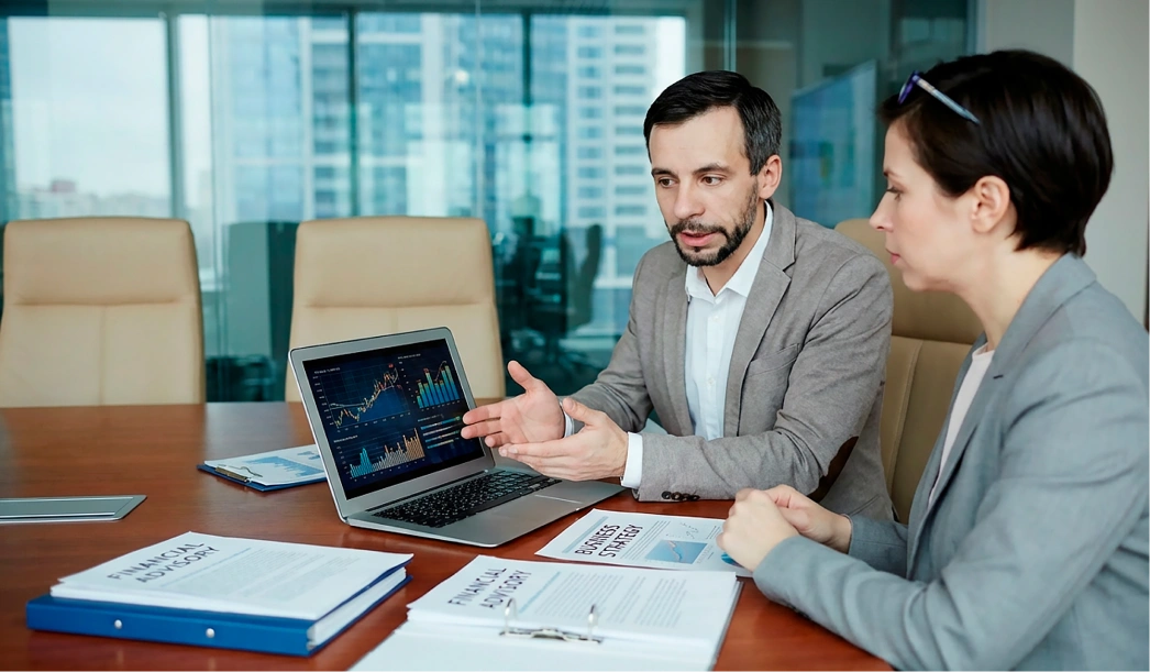 Two business professionals in a meeting reviewing financial charts on a laptop and documents on a conference table.