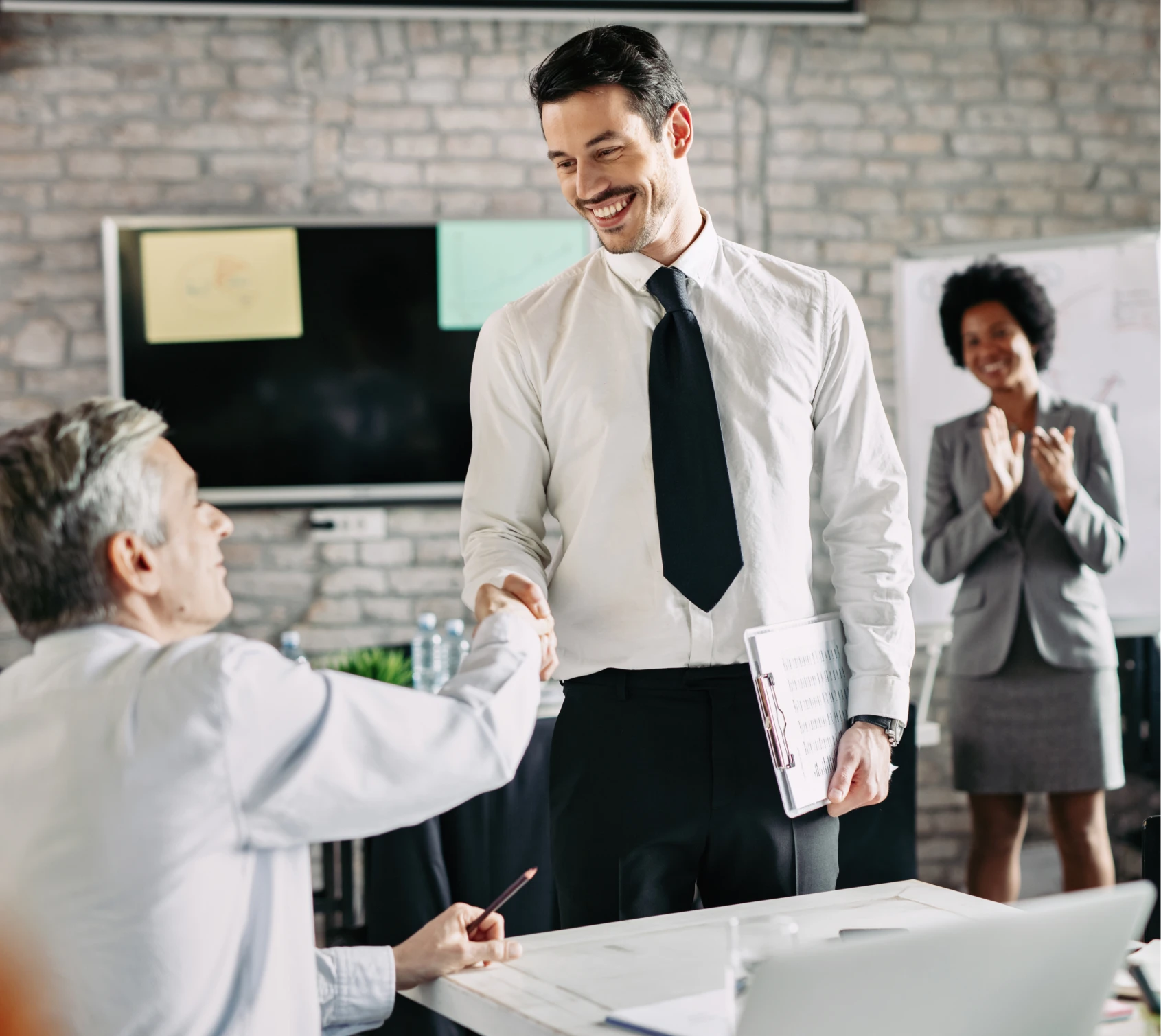 Smiling businessman shaking hands with a seated colleague while a woman claps in the background during a meeting.