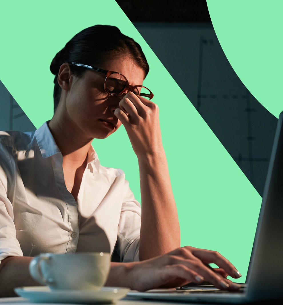 A woman wearing glasses and a white shirt sits at a laptop, pinching the bridge of her nose as if stressed or tired, with a coffee cup in the foreground.