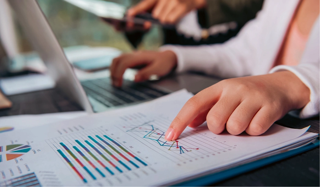 Person pointing at a line graph on printed data charts beside a laptop where another person types.