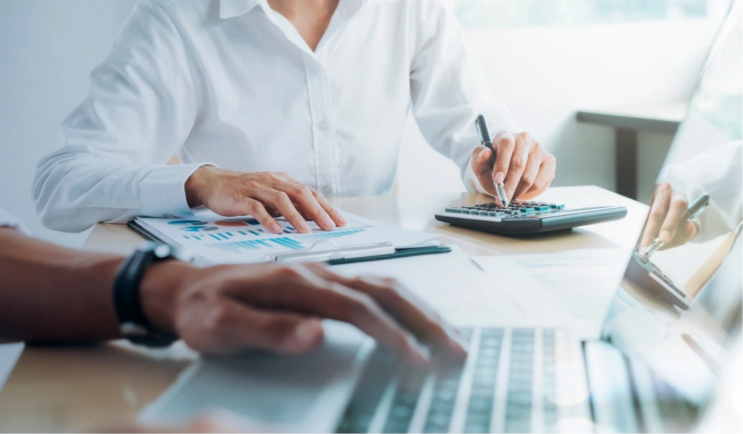 Person in white shirt using a calculator while another types on a laptop with financial documents on the table.