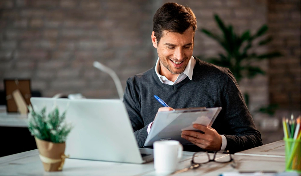 Smiling man in a dark sweater writing on a clipboard at a desk with a laptop, coffee mug, and potted plant.