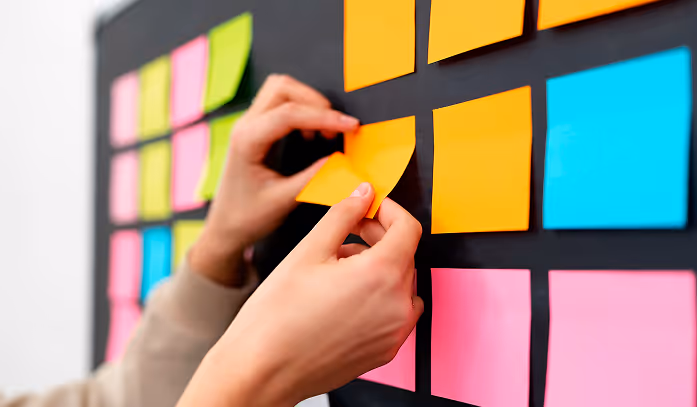 Person placing orange sticky notes on a black wall covered with colorful sticky notes in green, pink, and blue.