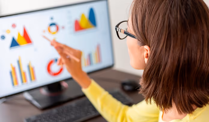 Person wearing glasses pointing at colorful graphs and charts on a computer screen.