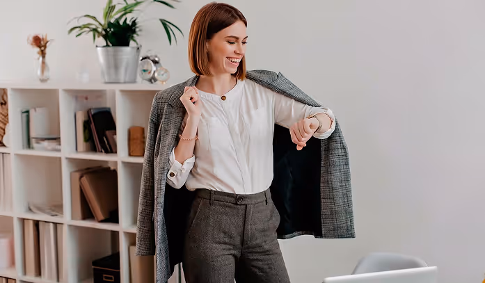 Smiling woman wearing gray pants and white blouse, putting on a gray blazer and checking her wristwatch in a modern office.