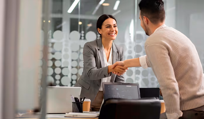 Two business professionals smiling and shaking hands across a desk in a modern office.