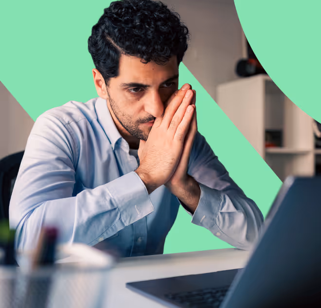 Man with curly hair in a light blue shirt looking intently at a laptop with hands pressed together near his face.