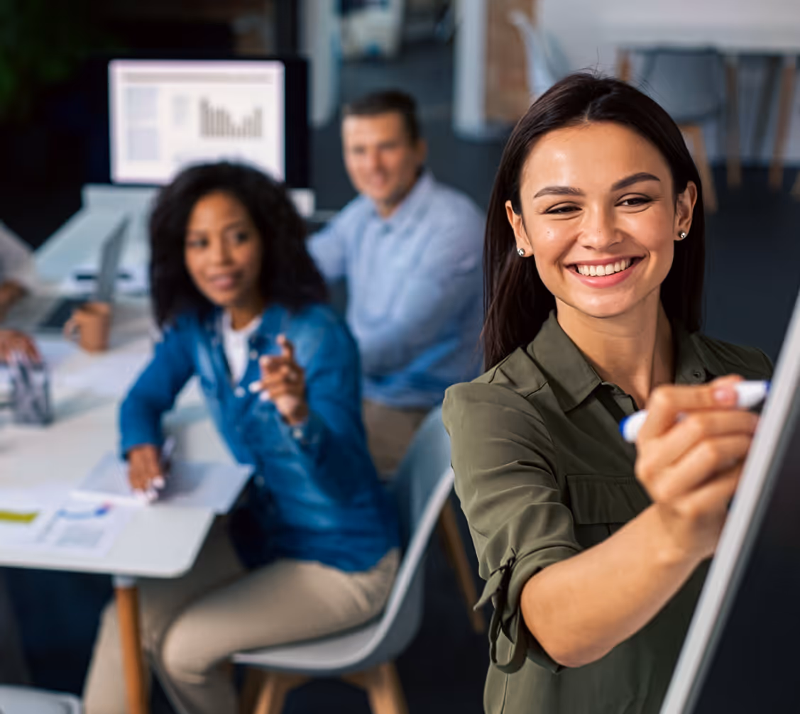 Smiling woman writing on a whiteboard during a meeting with colleagues in an office.