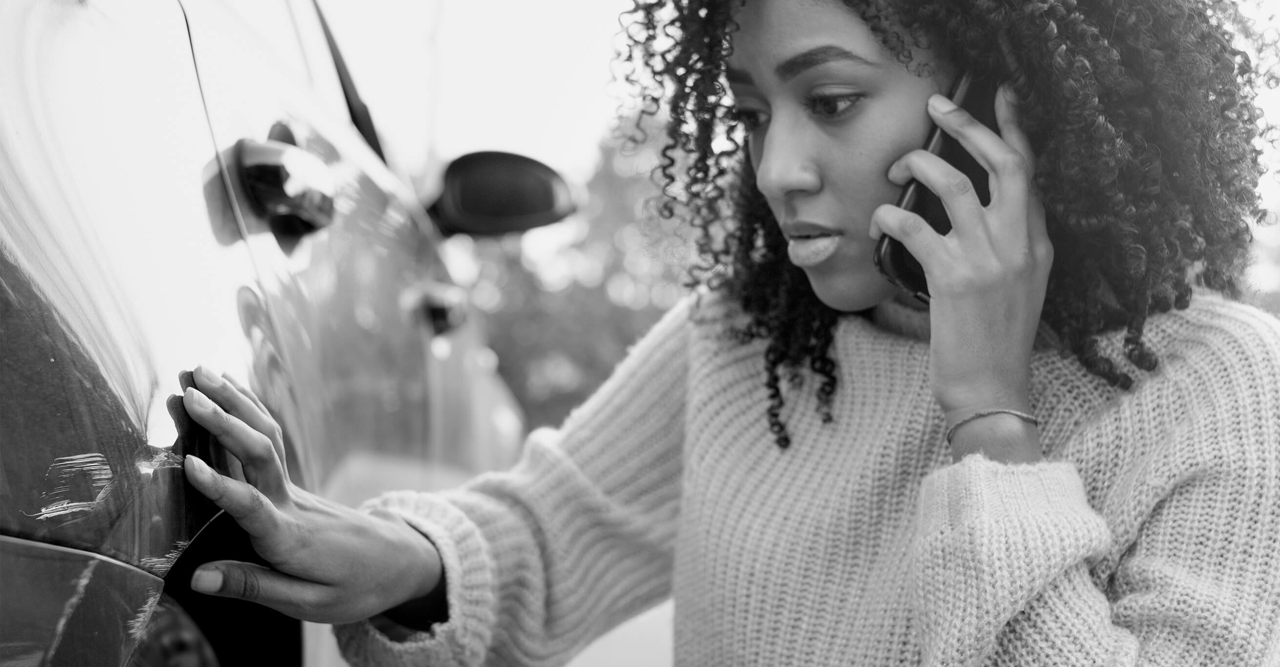 A woman inspects her car while calling her insurance company to find out who pays for car damage in a no-fault state.
