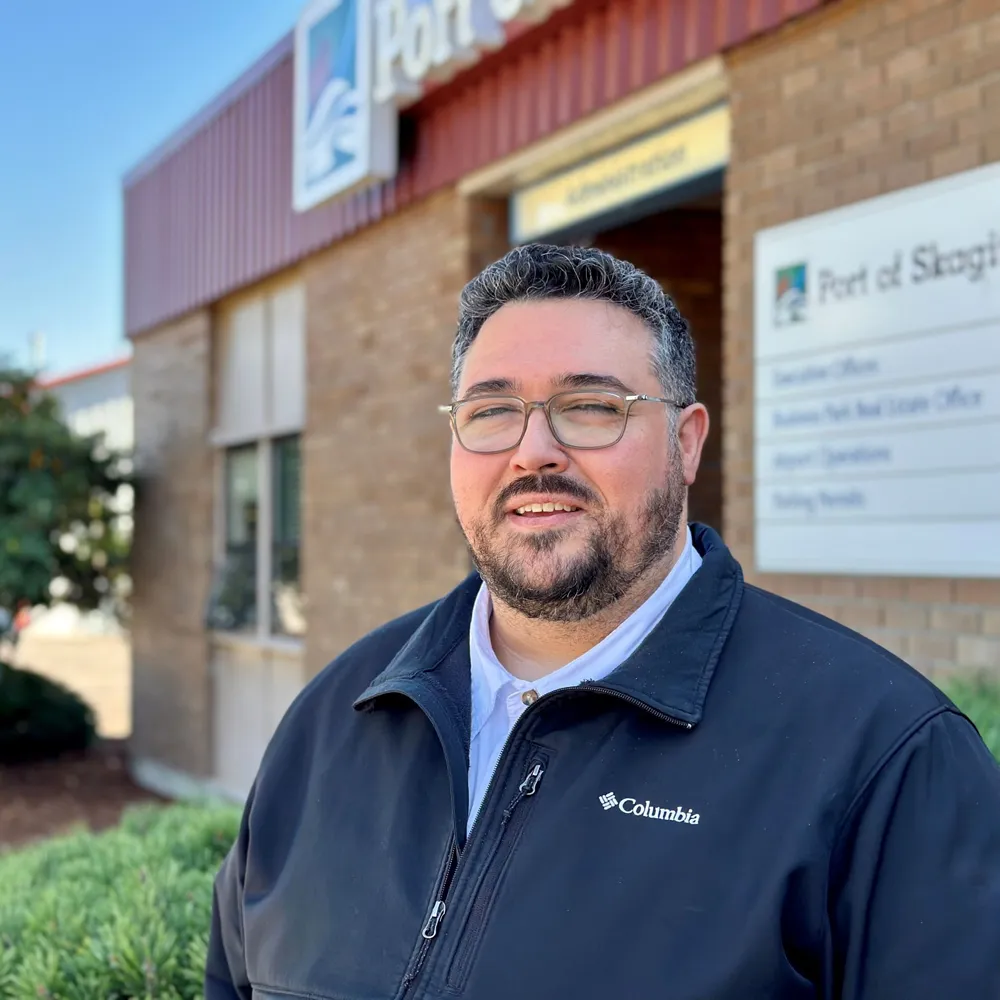 Man with glasses and beard wearing a black Columbia jacket standing outside a brick building with a Port of Skegee sign.