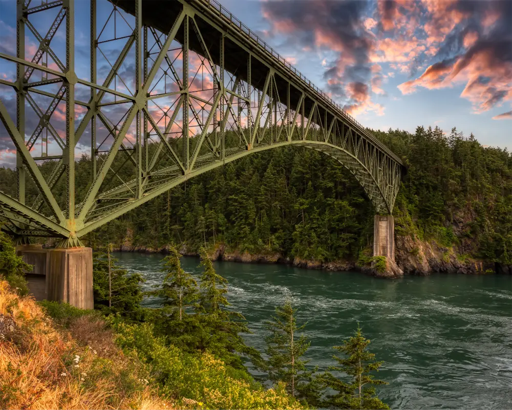 Large steel arch bridge crossing a river with dense evergreen forest on the riverbanks under a dramatic sunset sky.