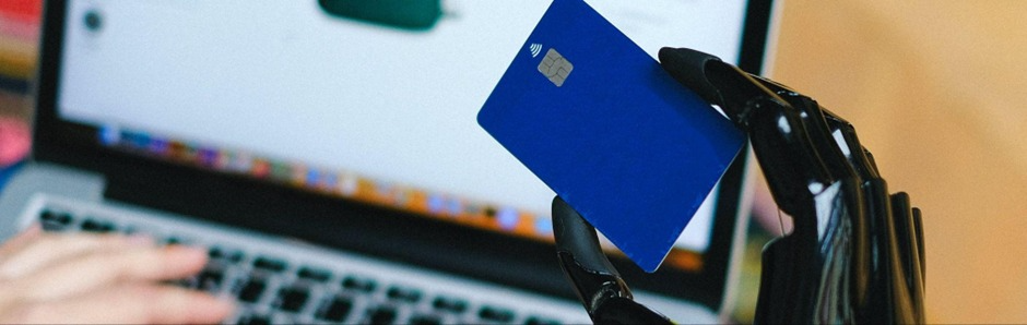A robotic hand holds a blue credit card in front of a laptop while a human hand types on the keyboard.