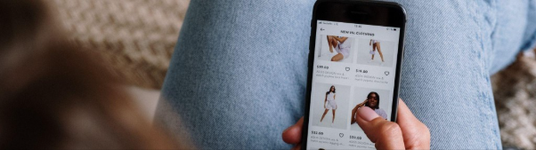 A person holds a smartphone and browses an online clothing store, viewing various women’s outfits and prices while sitting cross-legged in jeans.