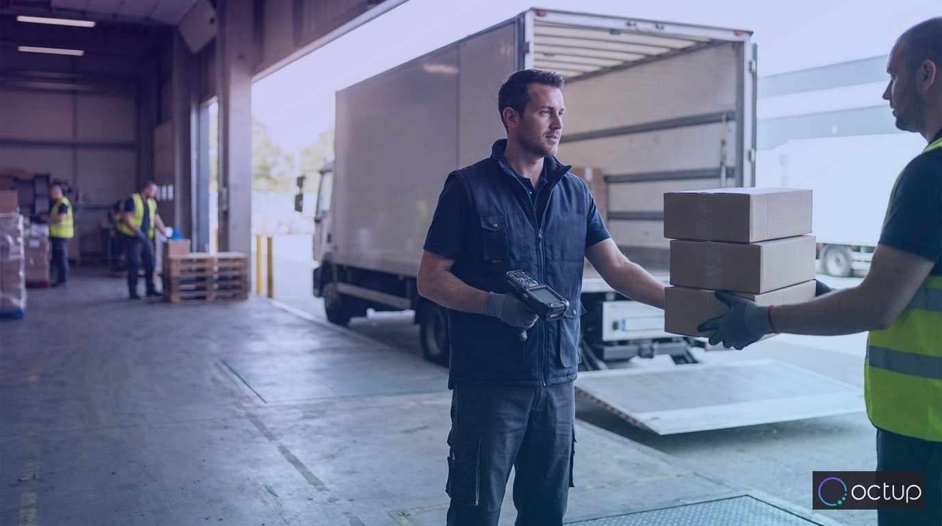 A uniformed carrier driver accepting parcels from a dock worker at a warehouse loading dock beside an open delivery truck.