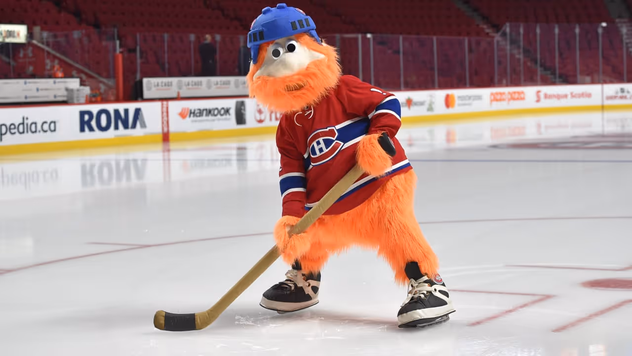 The Montreal Canadiens' orange furry mascot wearing a blue helmet and the team's red jersey holds a hockey stick on the ice rink.