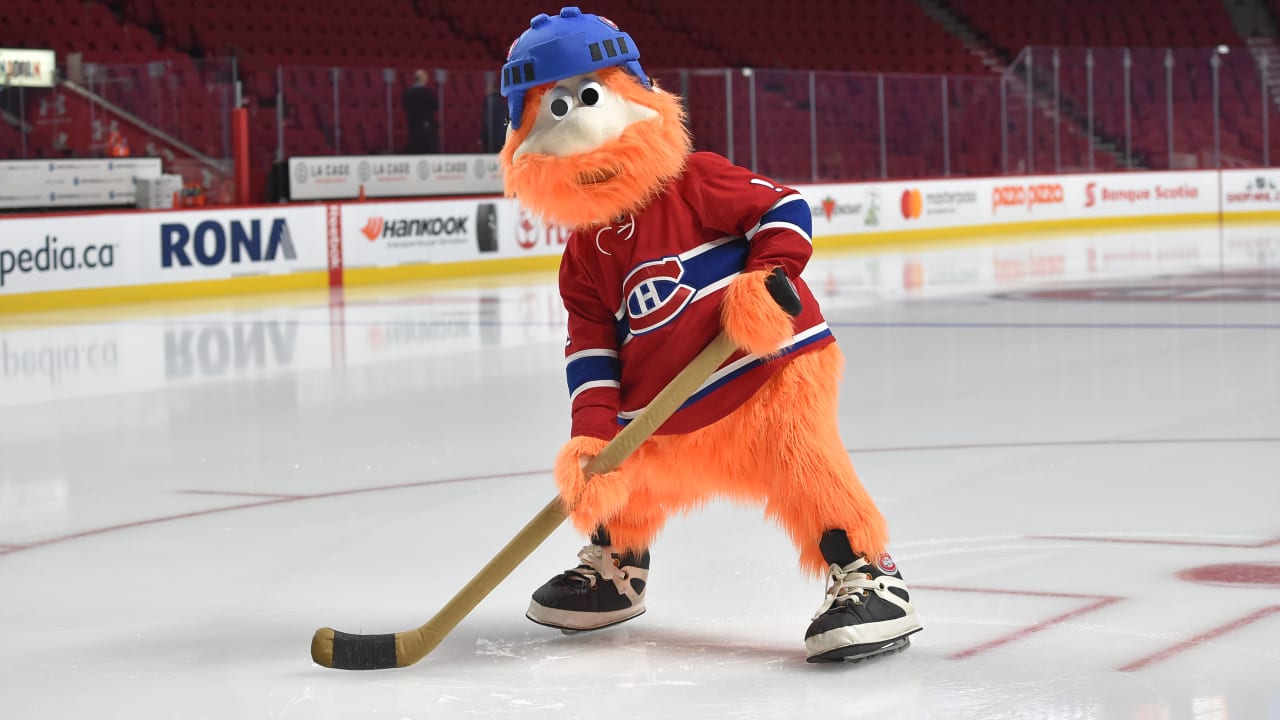 The Montreal Canadiens' orange furry mascot wearing a blue helmet and the team's red jersey holds a hockey stick on the ice rink.