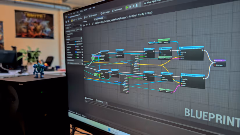 Two people working at a desk with dual monitors displaying game development software and simulation blueprints.