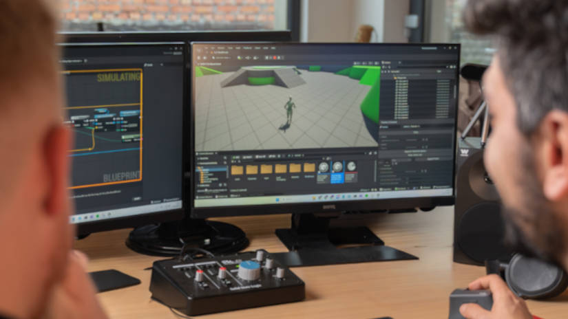 Two people working at a desk with dual monitors displaying game development software and simulation blueprints.