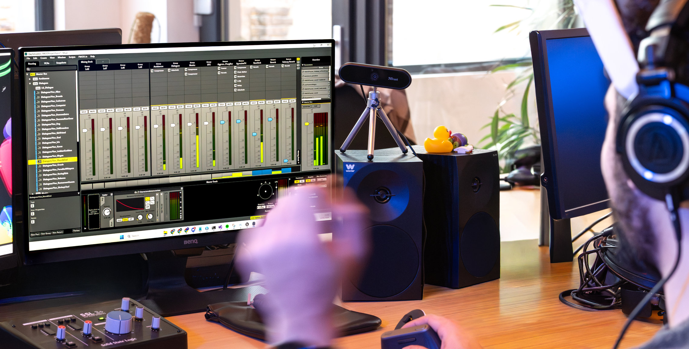 Person wearing headphones working on audio mixing software displayed on a desktop monitor with speakers and a webcam on the desk.