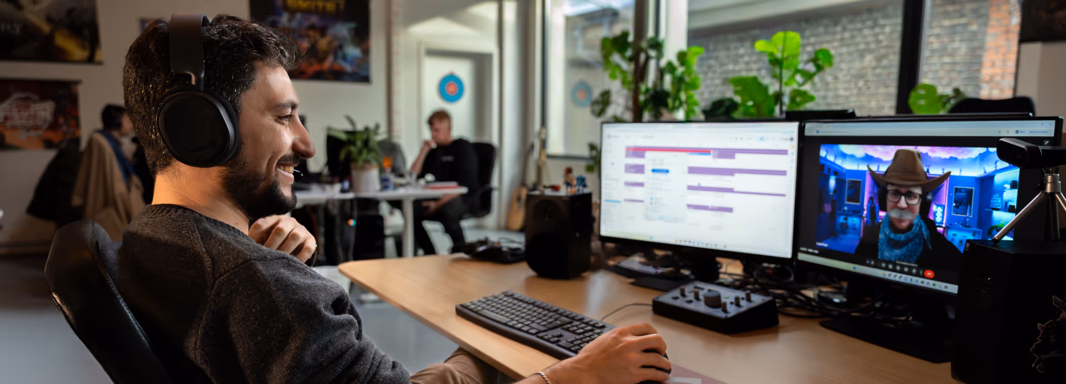 Man wearing headphones, smiling while working at a desk with dual monitors, one showing a video call with a cowboy hat filter.