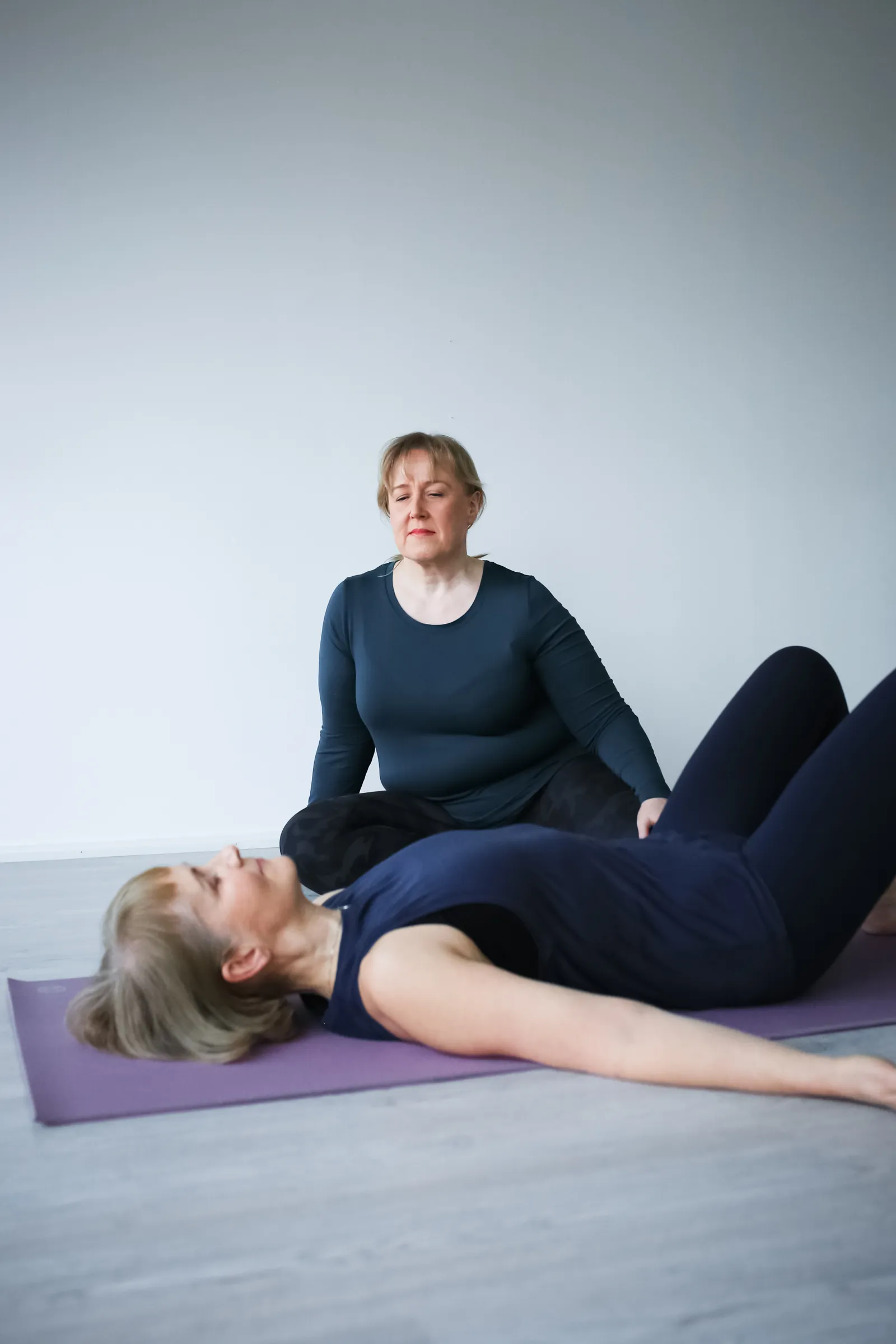 Kaksi naista joogaamassa vaaleassa huoneessa, toinen makaa matolla ja toinen istuu ristijalkaisesti.