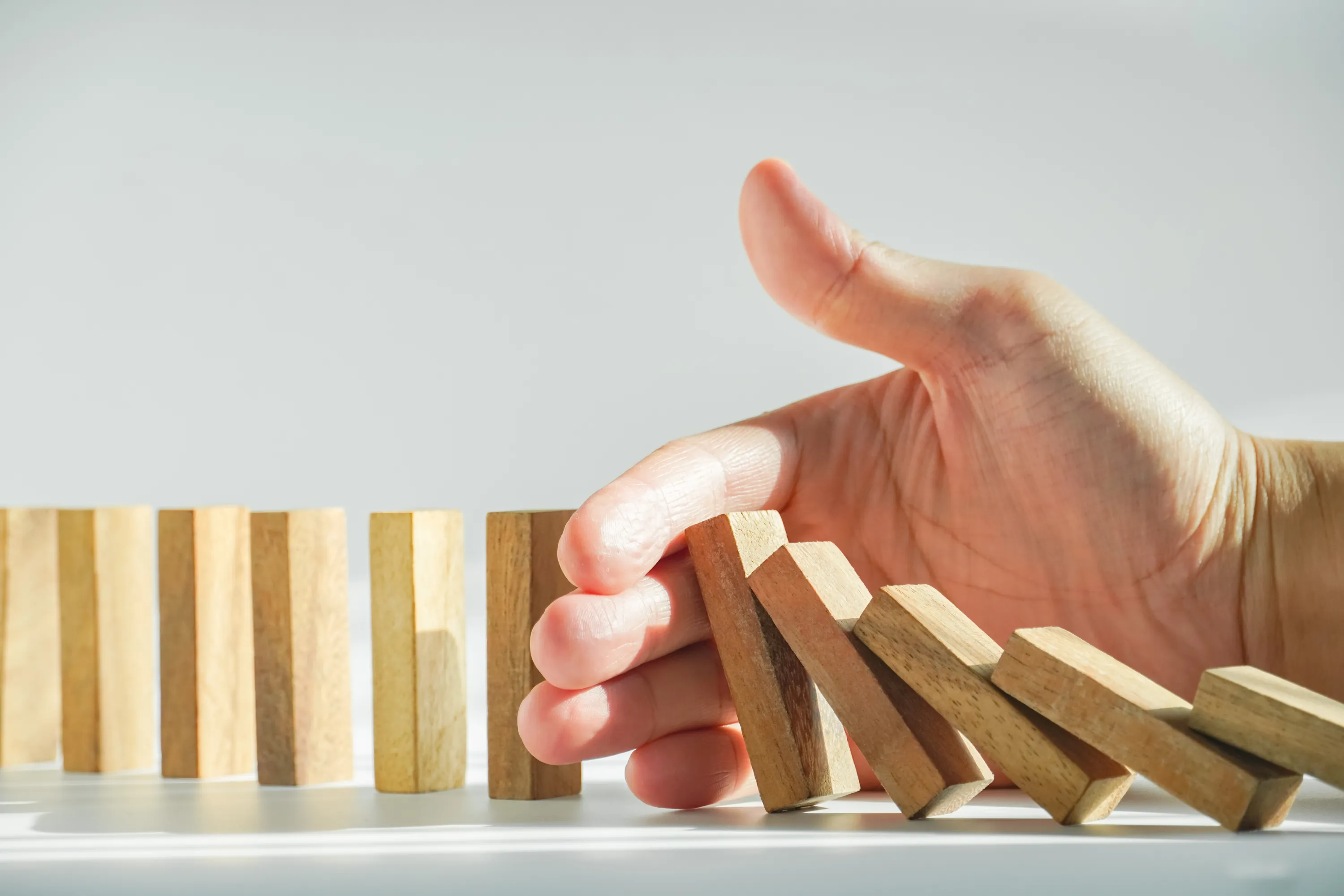 Käsivarren pysäyttämät puiset dominopalikat valkoisella taustalla, valaistus on kirkas.