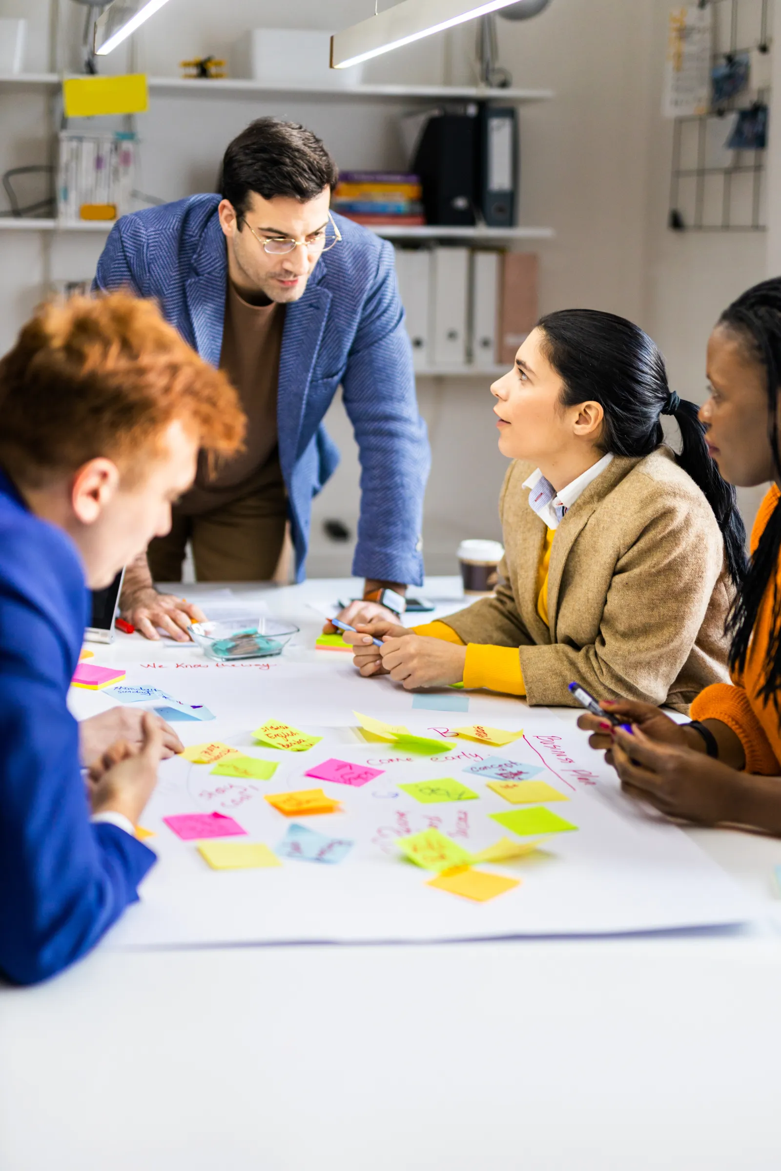 Neljä ihmistä työskentelee yhdessä pöydän ääressä, jossa on värikkäitä muistilappuja, kirkas valaistus.
