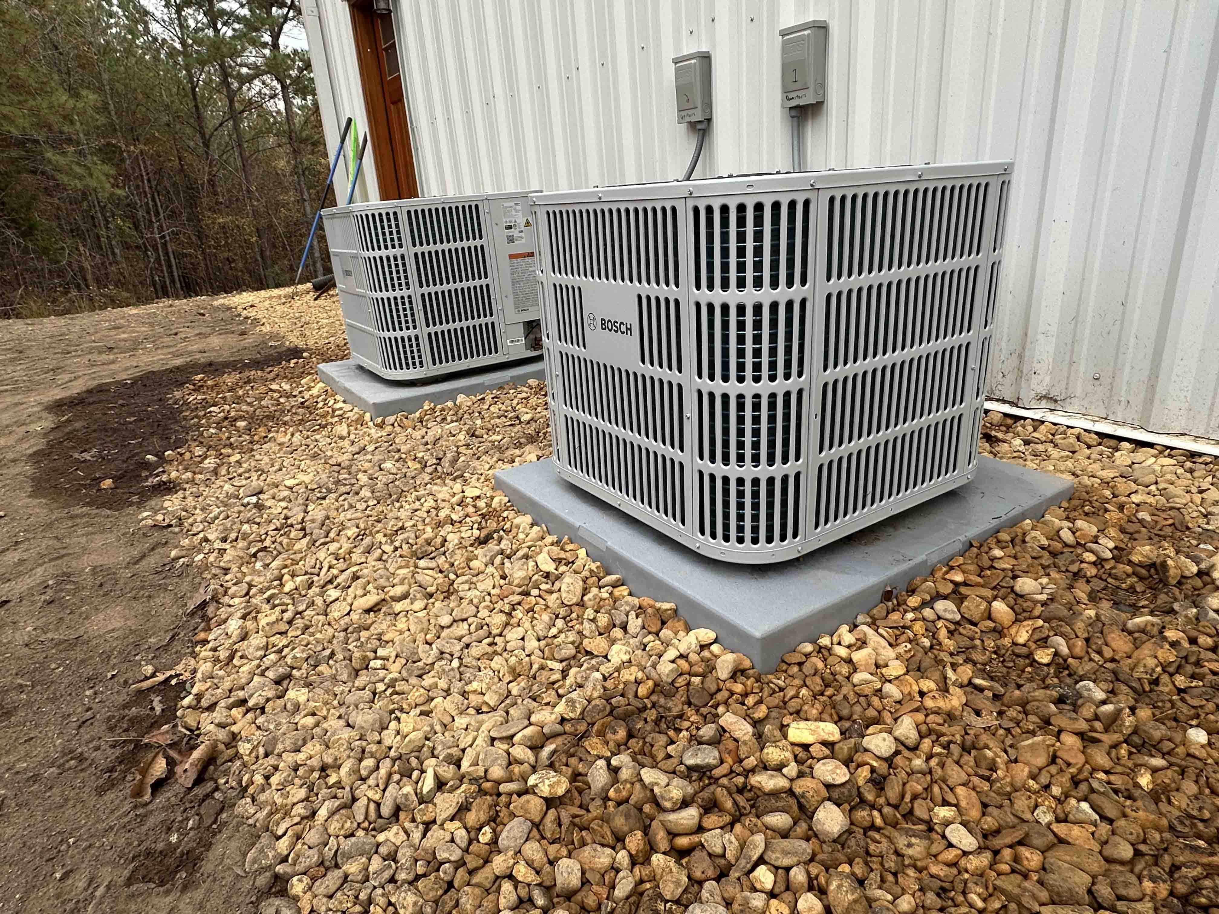 Two gray Bosch HVAC units installed on concrete pads next to a white metal building surrounded by small brown stones.