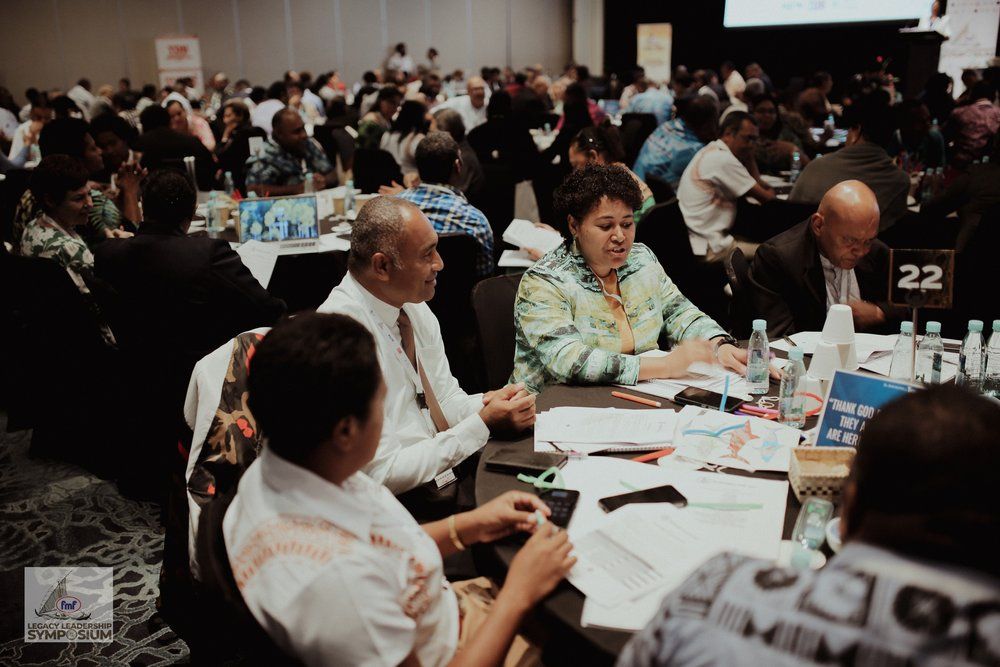 A group of three men collaborate at a wooden table in a modern, open-plan office. Laptops and soda cans are scattered around. The atmosphere is focused and collaborative.