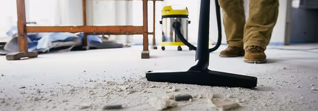 Vacuum cleaner nozzle on a dusty floor with construction debris, a person's legs and workbench in the background.