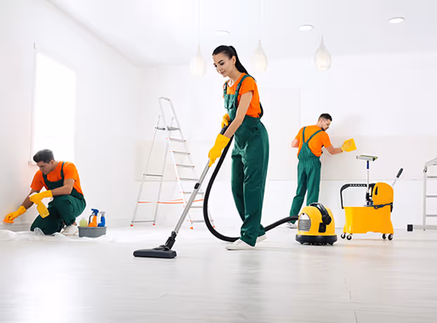 Three workers in orange shirts and green overalls cleaning a bright white room, one vacuuming the floor, one scrubbing the wall, and one wiping near the window.
