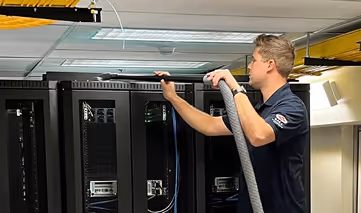 Technician cleaning or inspecting ceiling ventilation above data center server racks using a vacuum hose.