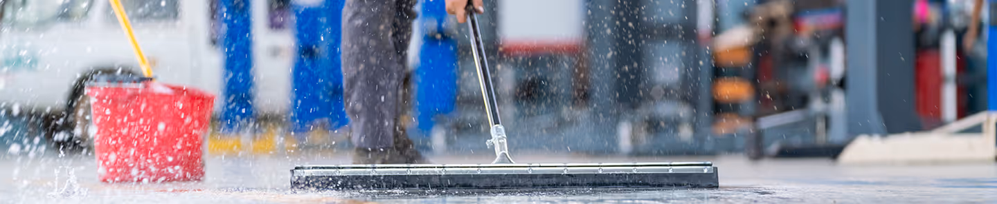 Close-up of a person cleaning a wet floor with a squeegee, water splashing, and a red bucket nearby in a workshop.