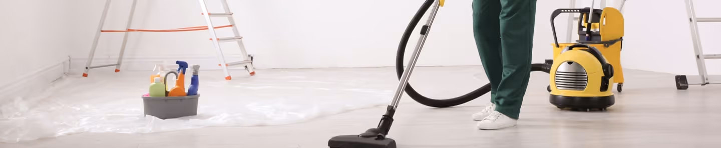 Person vacuuming a floor in a clean, bright room with cleaning supplies and ladders in the background.