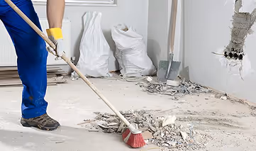 Person in work boots and gloves sweeping dust and debris on a construction site floor with a broom.