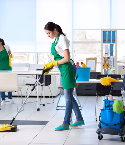 Woman in a green apron and yellow gloves cleaning a tiled floor with a vacuum cleaner in a bright office.