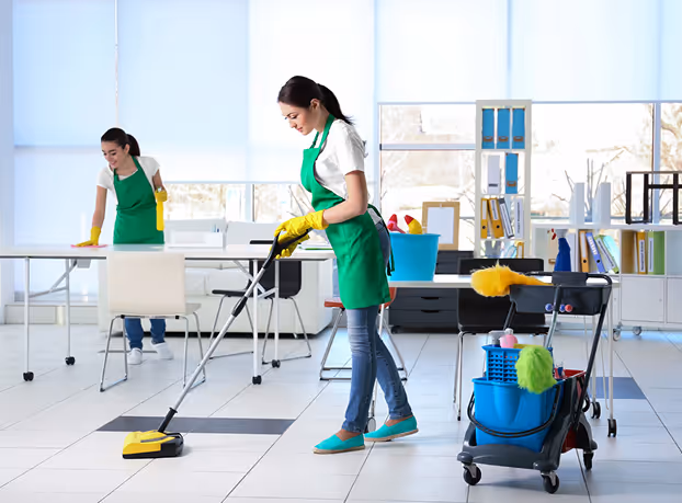 Two women wearing green aprons and yellow gloves cleaning an office with a vacuum and spray bottles.