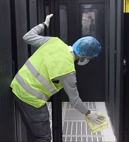 Person wearing a yellow safety vest, blue hair cover, face mask, and gloves cleaning the floor inside a data center aisle.