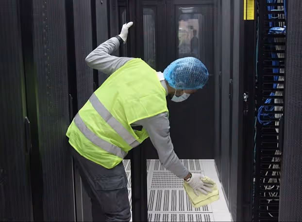 Worker wearing a neon yellow safety vest, blue hairnet, and white gloves cleaning the floor inside a server room.