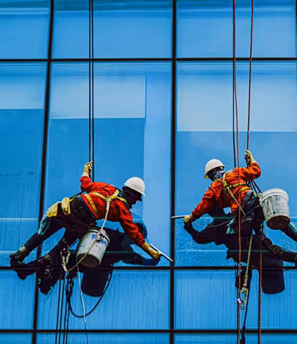 Two workers in orange uniforms and helmets suspended by ropes cleaning large glass windows of a tall building.