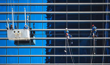 Workers cleaning the glass facade of a tall building, with two suspended by ropes and two standing on a platform.